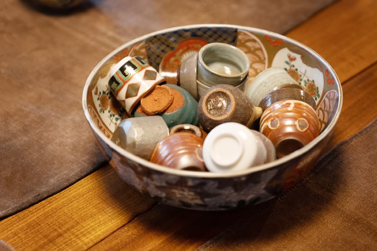 Tante piccole tazze da sake di ceramica giapponese, ognuna di forma e smalto diversi, raccolte dentro una ciotola decorata
