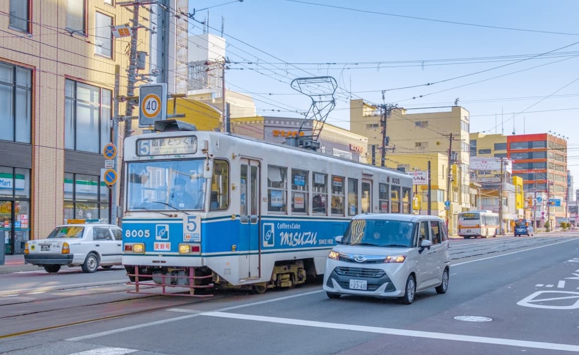 Un tram bianco e blu di Hakodate su una strada cittadina, con la destinazione in giapponese e i palazzi intorno