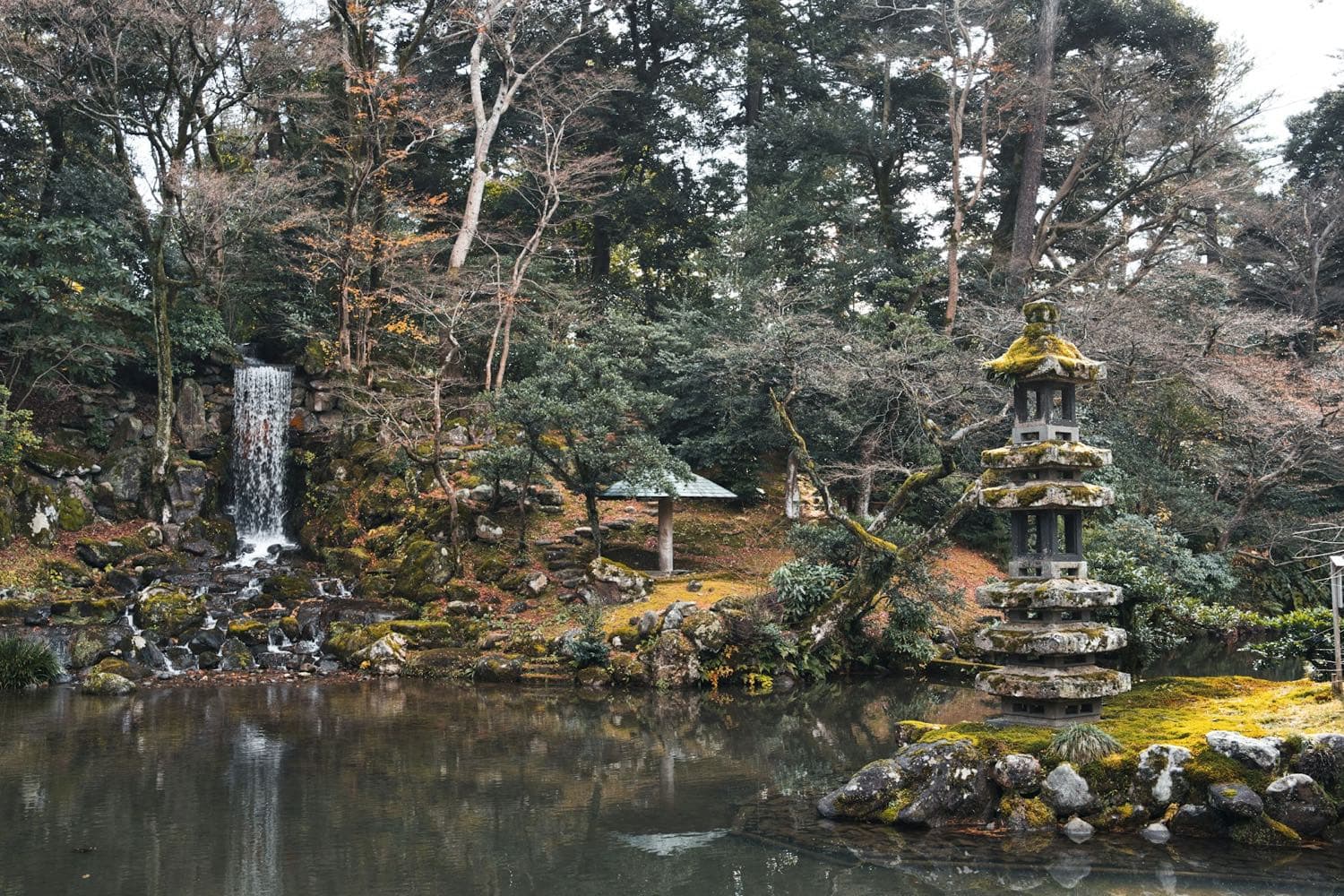 Uno stagno del giardino Kenroku-en con una piccola cascata, una pagoda di pietra ricoperta di muschio e gli alberi riflessi nell'acqua