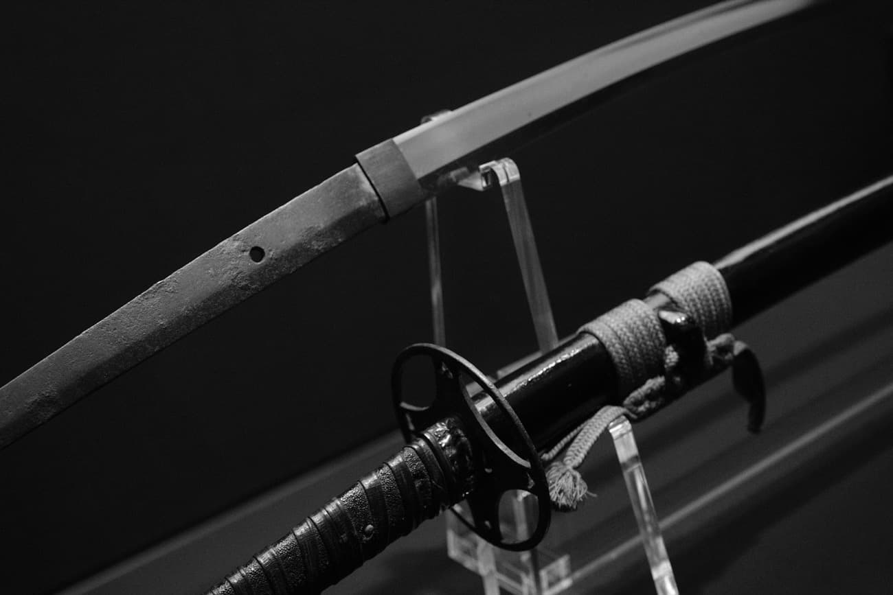 Una katana antica esposta su un supporto in un museo, con la lama nuda curva e l'elsa avvolta, in bianco e nero