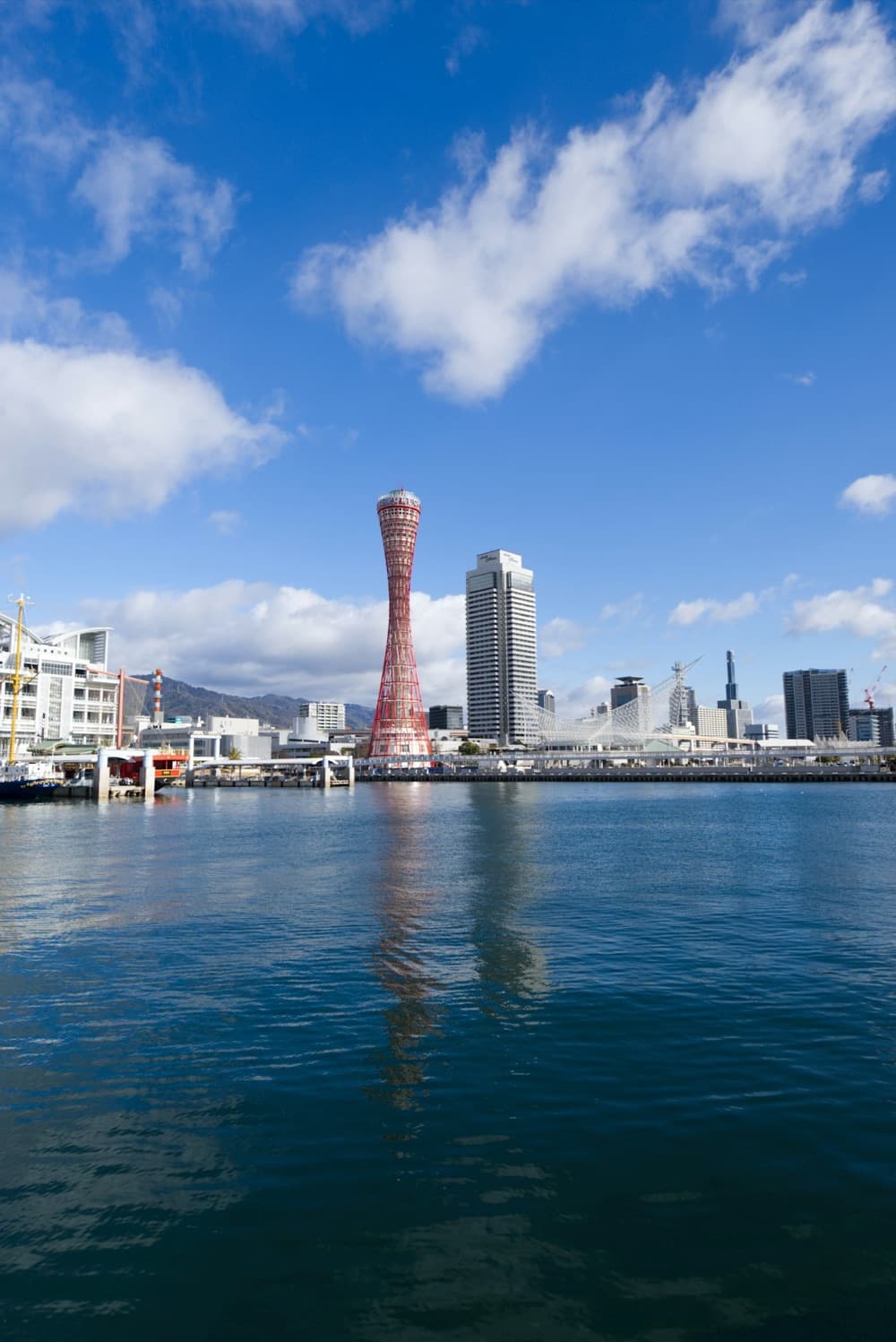 Il porto di Kobe con la torre rossa che si specchia sull'acqua, sotto un cielo azzurro