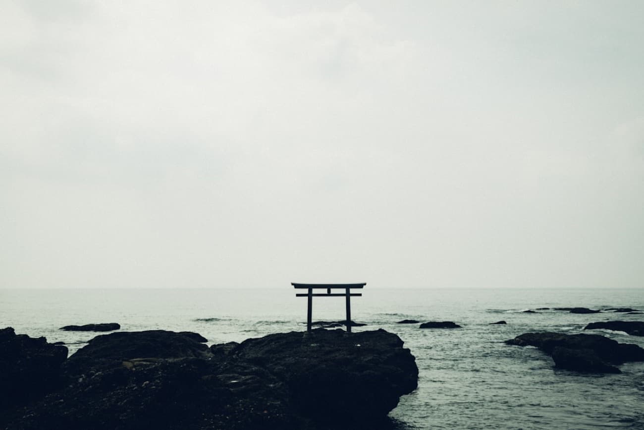 Un torii scuro che si erge tra gli scogli, in mezzo al mare, sotto un cielo velato