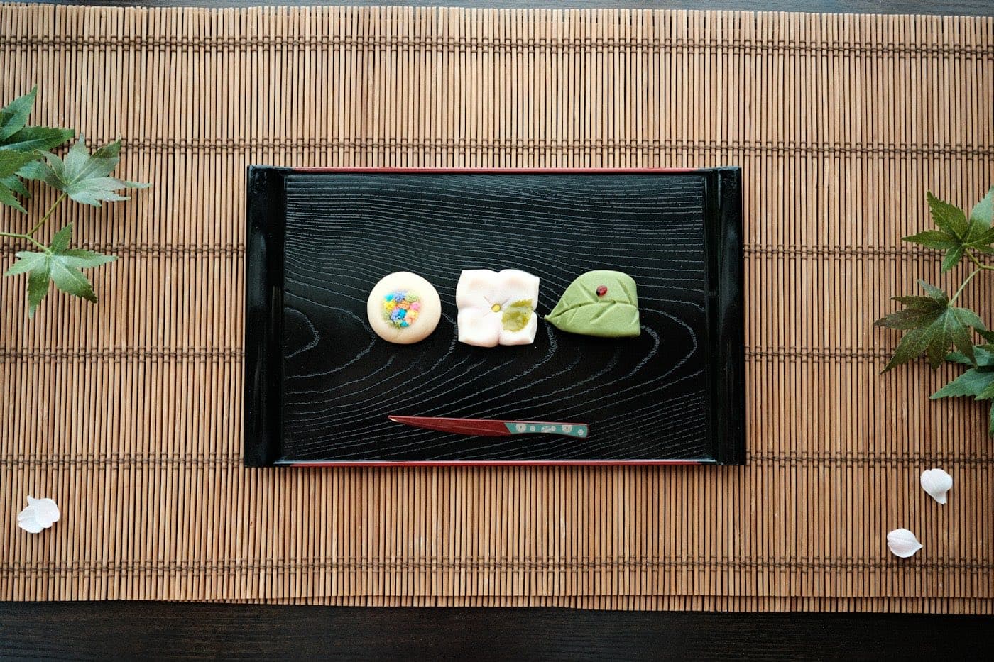 Tre wagashi di stagione su un vassoio laccato nero, con una forchettina, su una stuoia di bambù con foglie d'acero