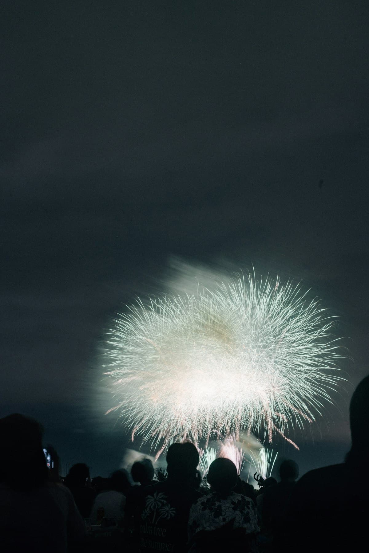 Un grande fiore di fuoco bianco esplode nel cielo notturno sopra la folla in silhouette a un festival estivo giapponese