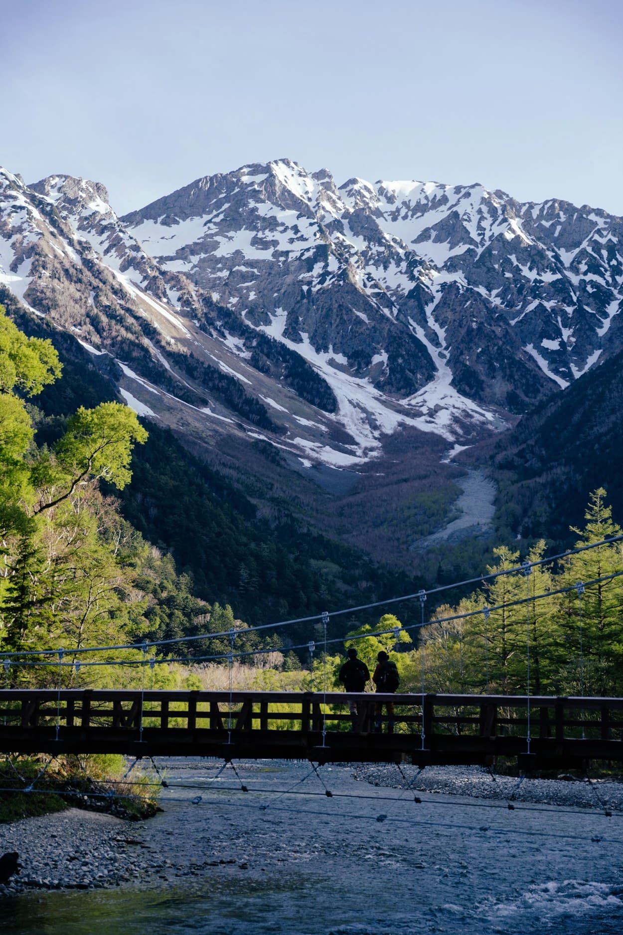 Il ponte Kappa di Kamikochi, un ponte sospeso di legno sul fiume, con le montagne innevate di Hotaka sullo sfondo