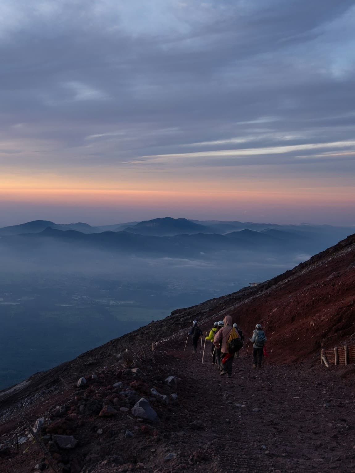 Escursionisti di spalle salgono il sentiero di terra vulcanica rossa del Monte Fuji all'alba, sopra un mare di nuvole