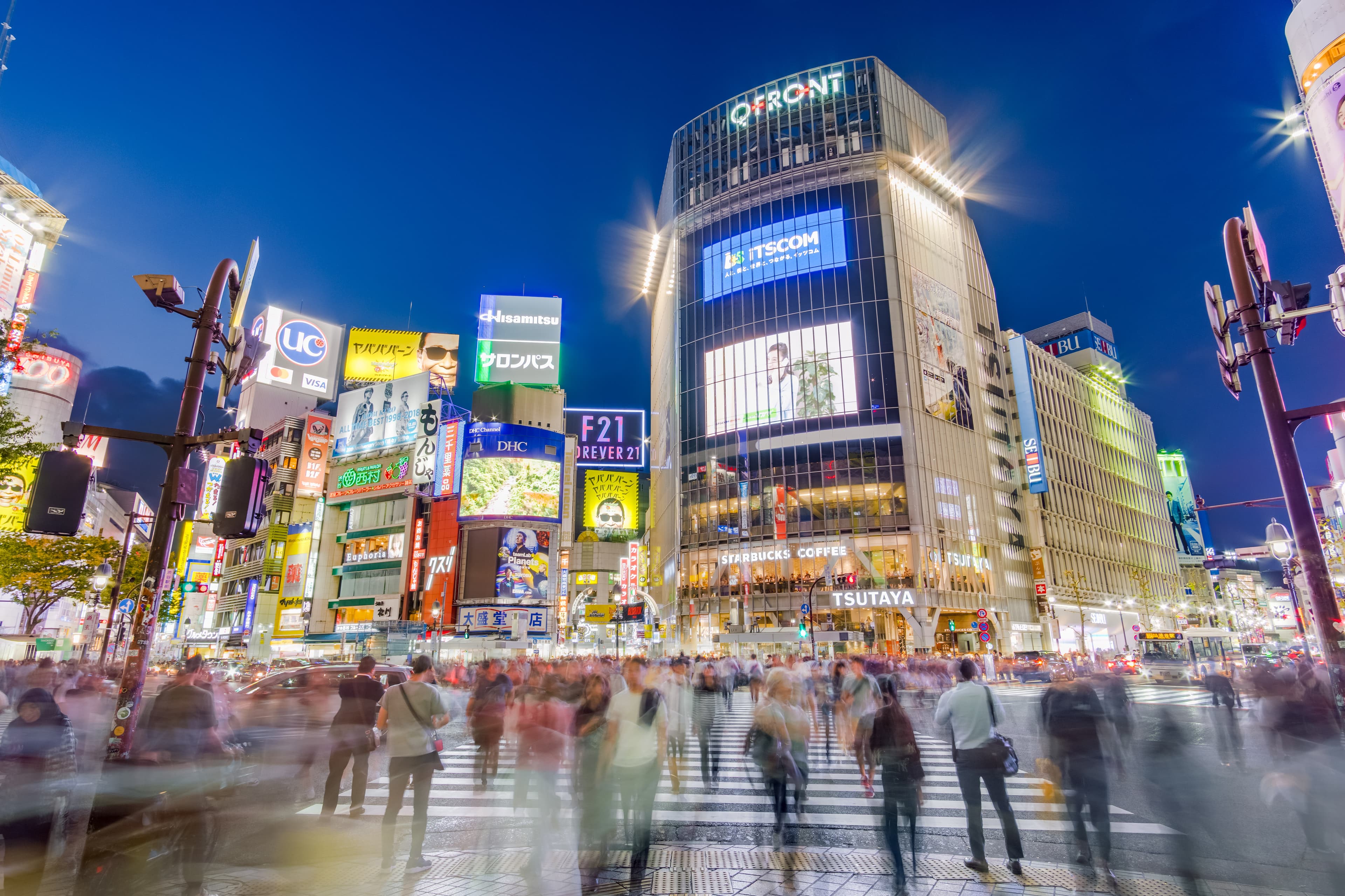 L'incrocio di Shibuya di notte, illuminato da insegne al neon e schermi giganti, con migliaia di persone che attraversano in tutte le direzioni