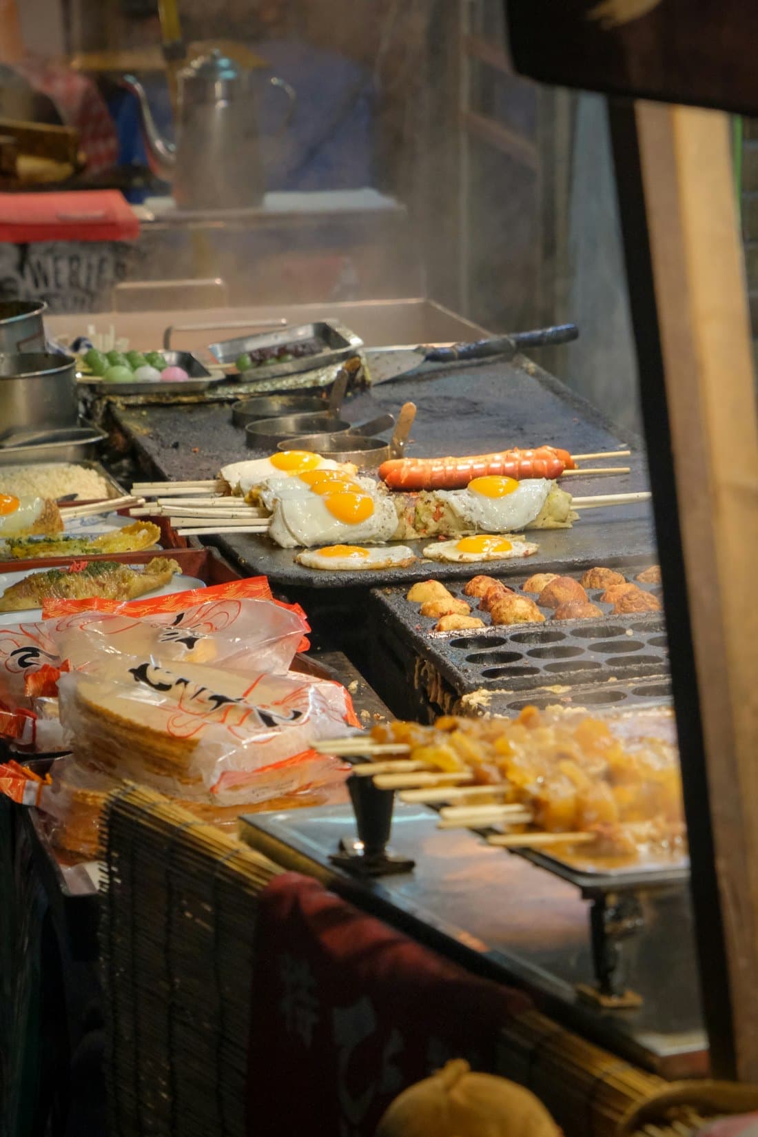Un banco di street food giapponese con una piastra per takoyaki, uova al tegamino, spiedini e dango colorati
