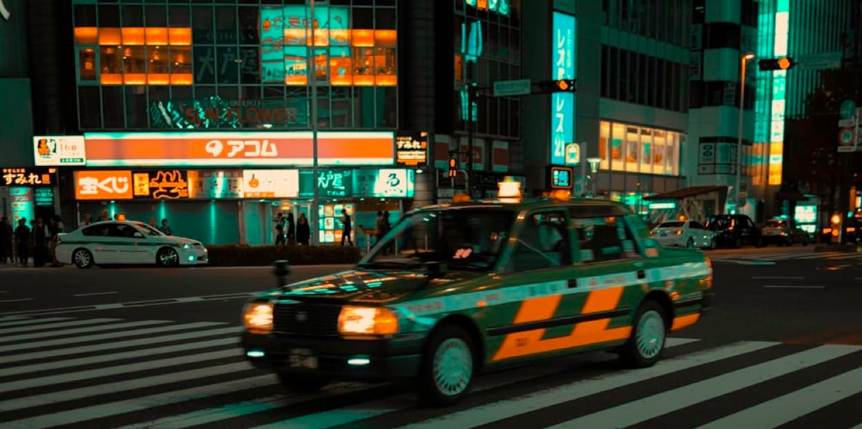 Un taxi giapponese verde e giallo attraversa un incrocio di notte, con le insegne luminose in giapponese sullo sfondo