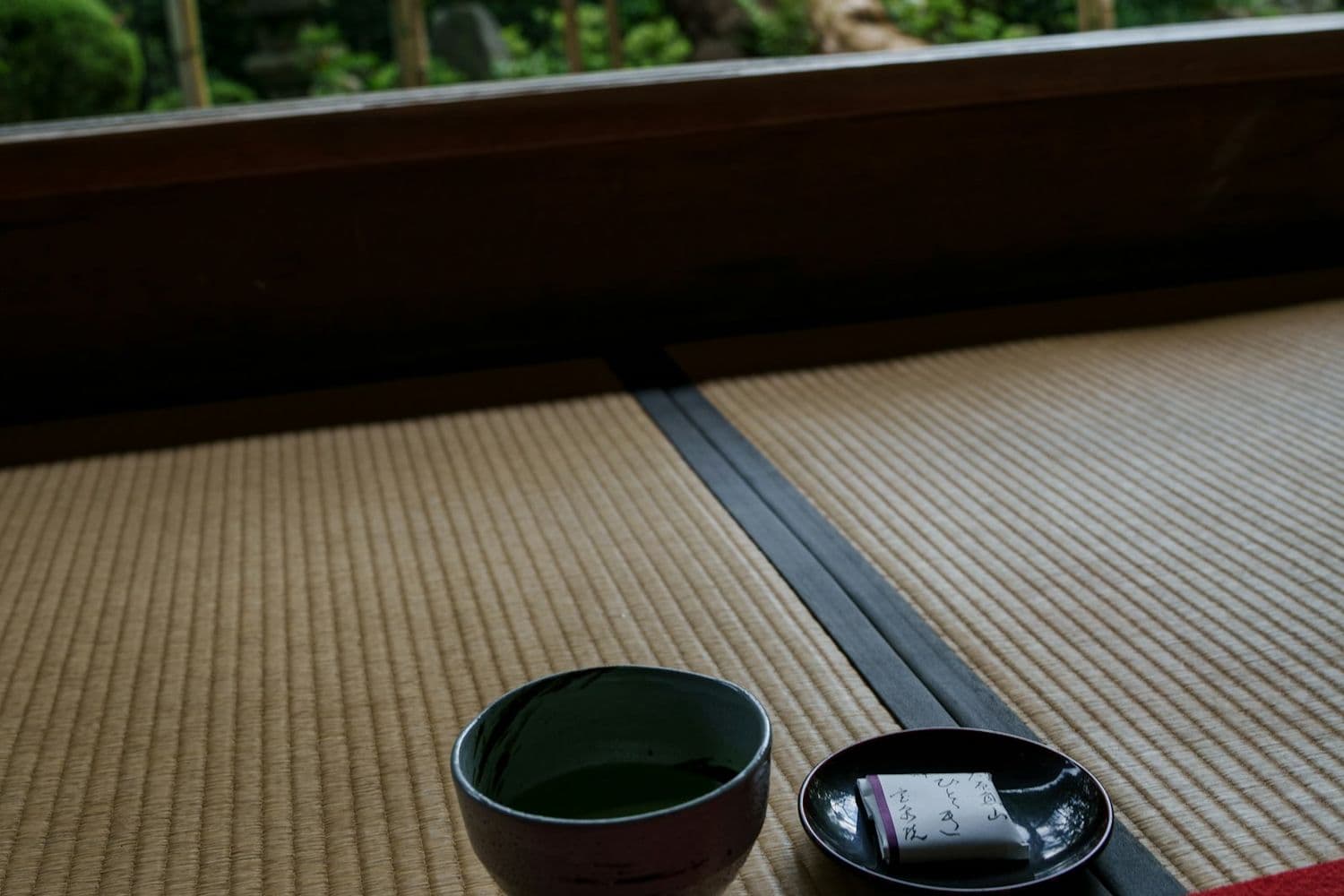 Una ciotola di tè matcha e un dolcetto su un piattino laccato, posati sul tatami davanti a un giardino verde