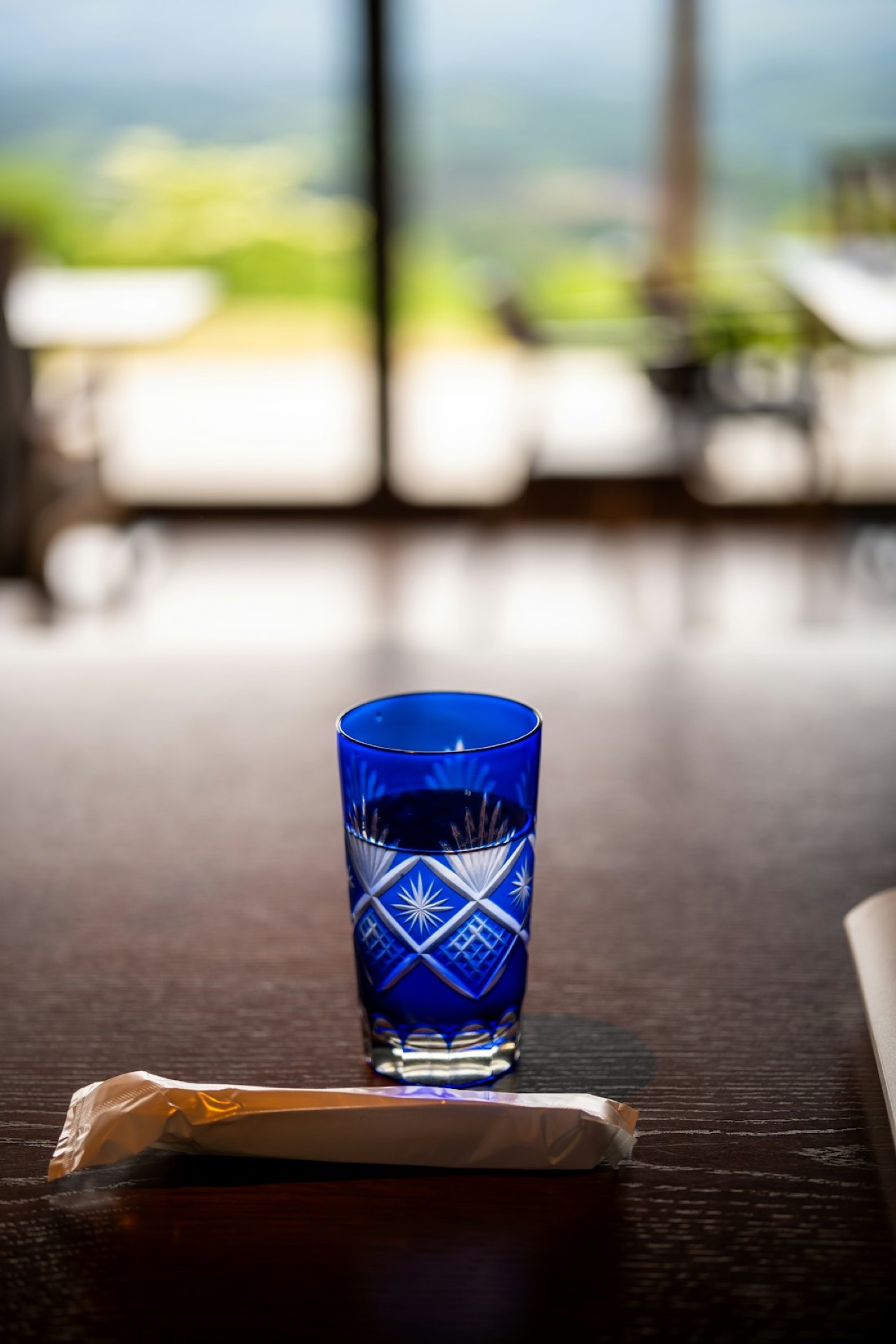 Un bicchiere di vetro tagliato Edo kiriko blu, con motivi geometrici finissimi, su un tavolo di legno davanti a una vista verde