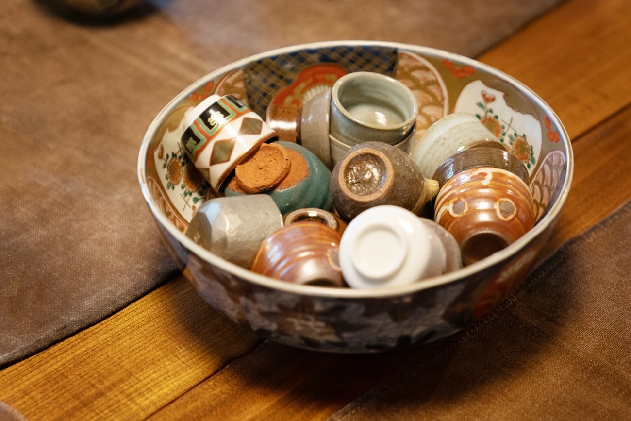 Una ciotola piena di tante piccole coppe di ceramica giapponese di stili e colori diversi, su un tavolo di legno