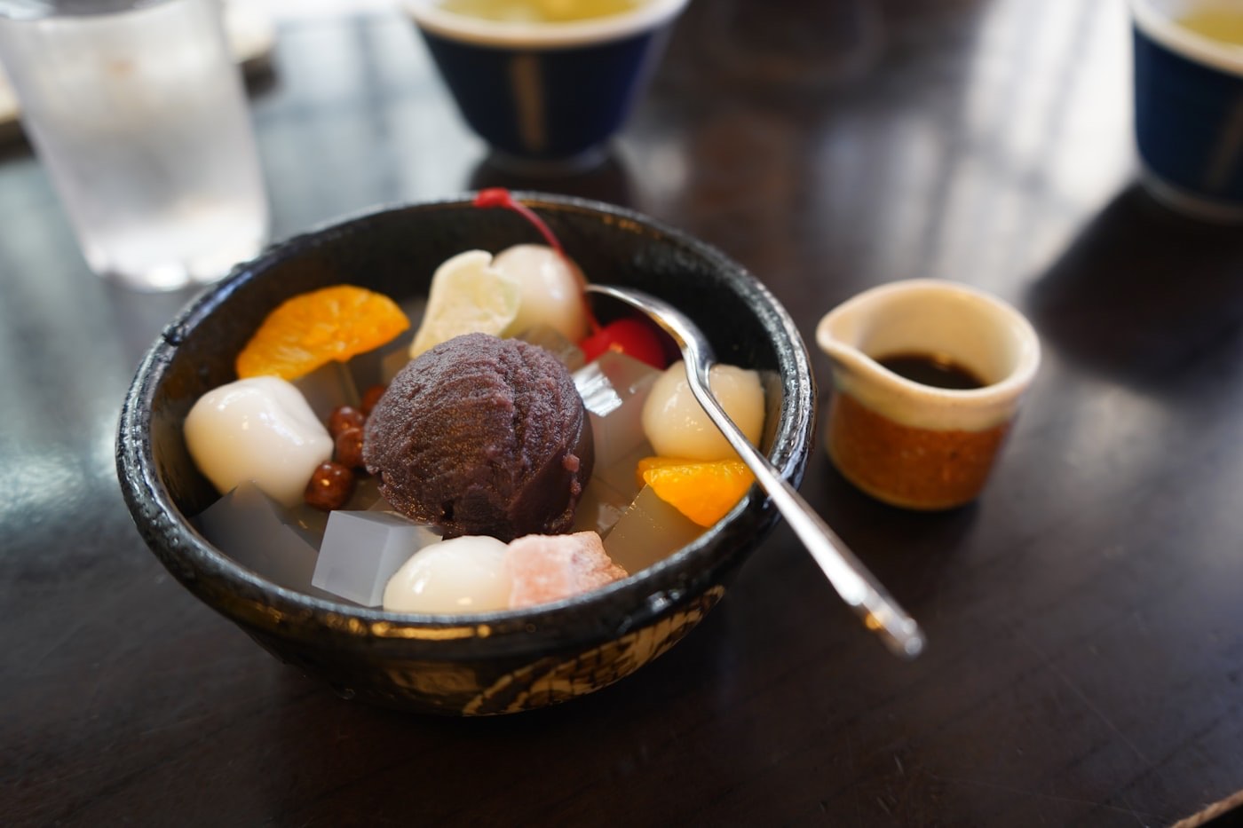 Una coppa di anmitsu: gelatina di kanten, mochi bianchi, frutta e una pallina di fagioli rossi, con lo sciroppo nero a parte