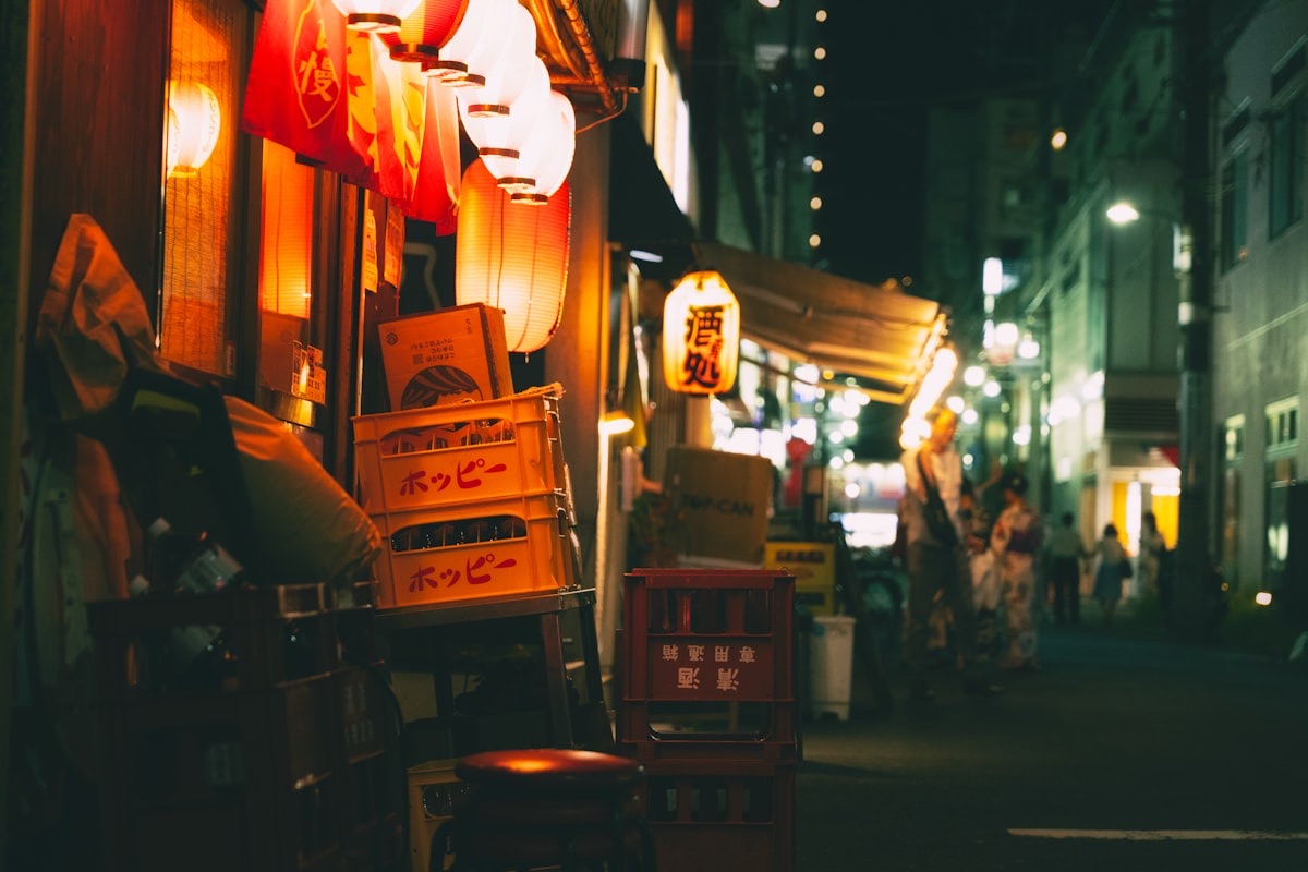 Una stradina di Tokyo la sera, con lanterne accese e casse di birra davanti a un'izakaya