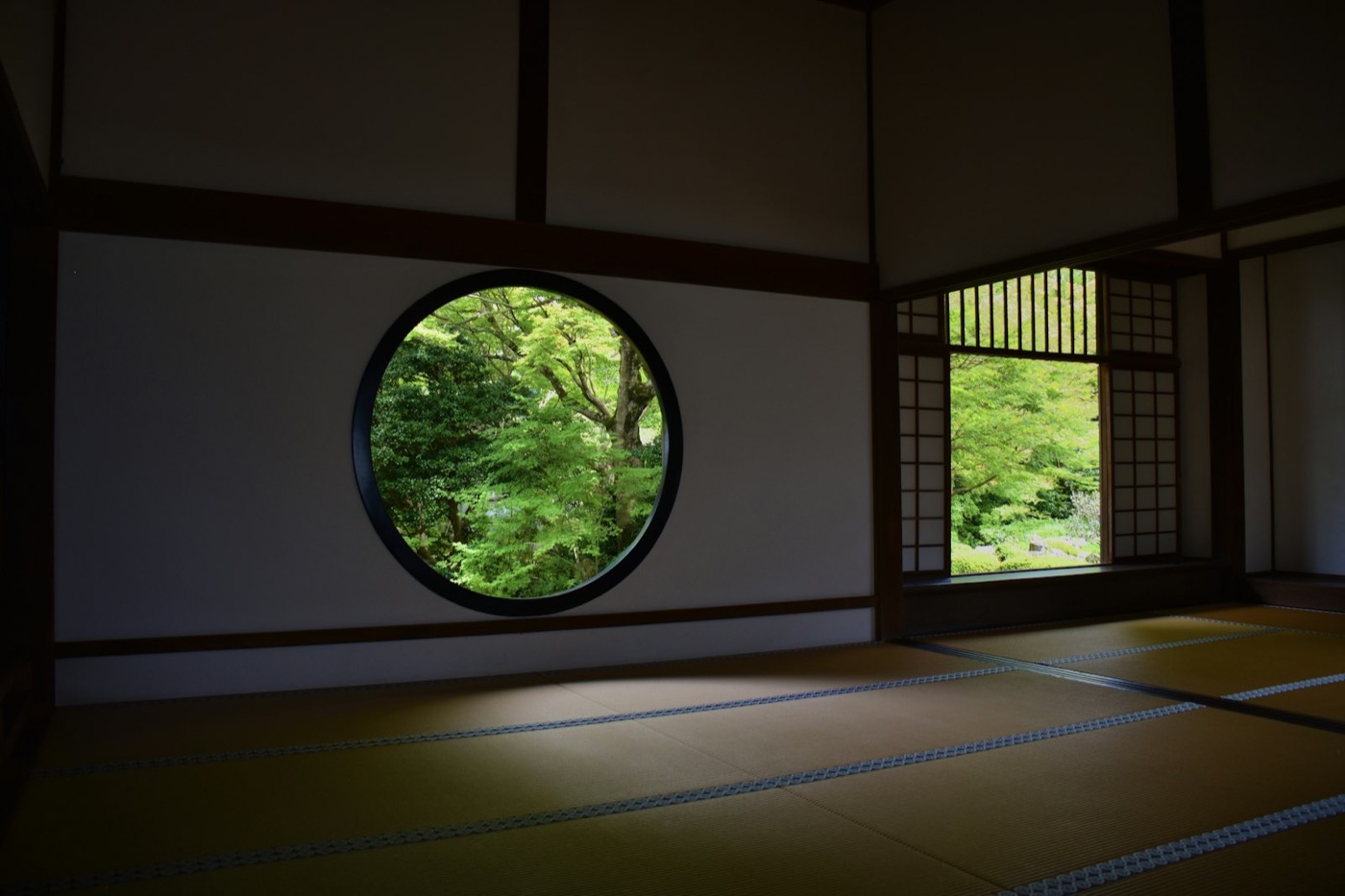 Una stanza di tatami con una grande finestra rotonda che inquadra il verde degli aceri del giardino
