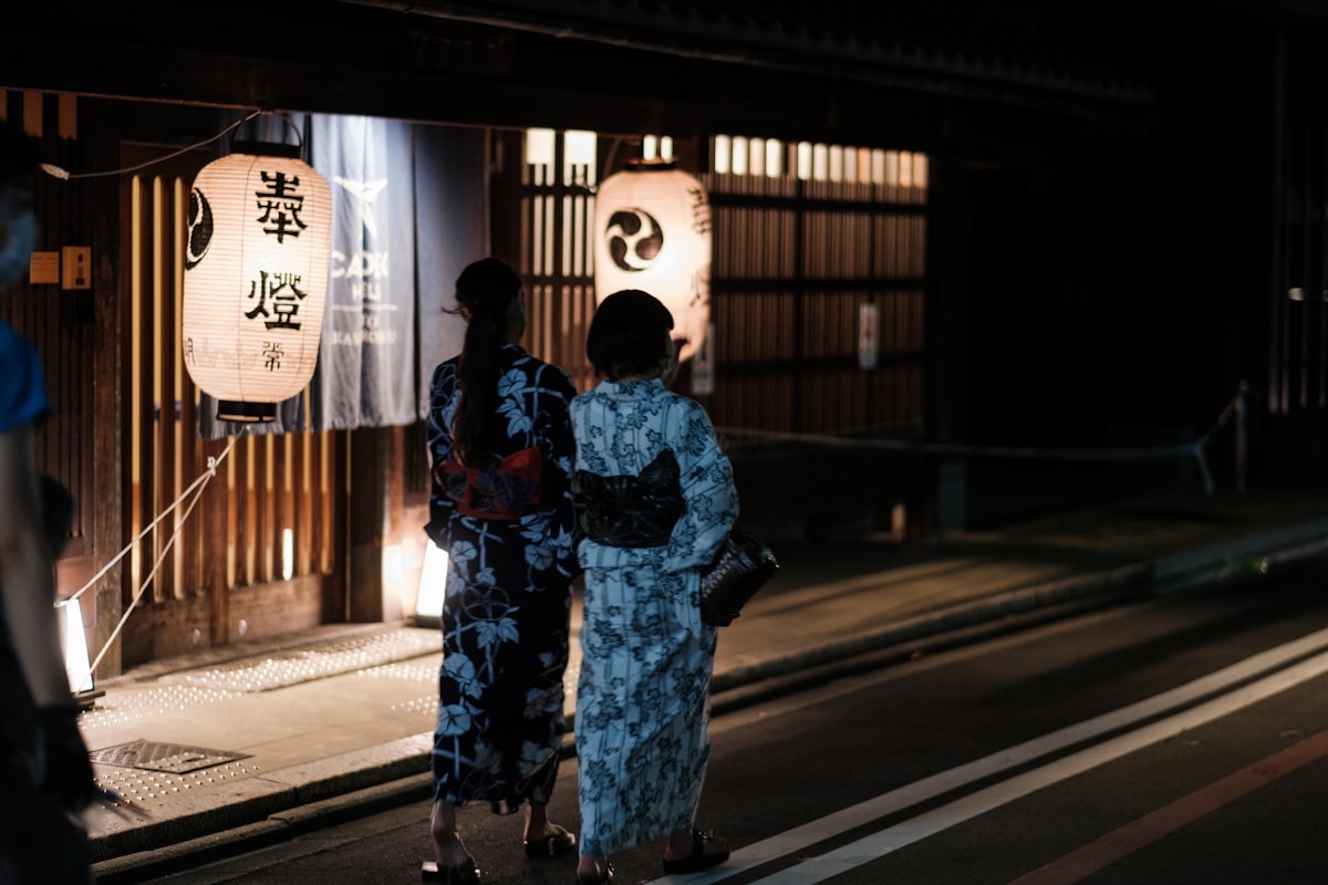 Due donne in yukata camminano la sera davanti alle lanterne di legno di un quartiere storico giapponese