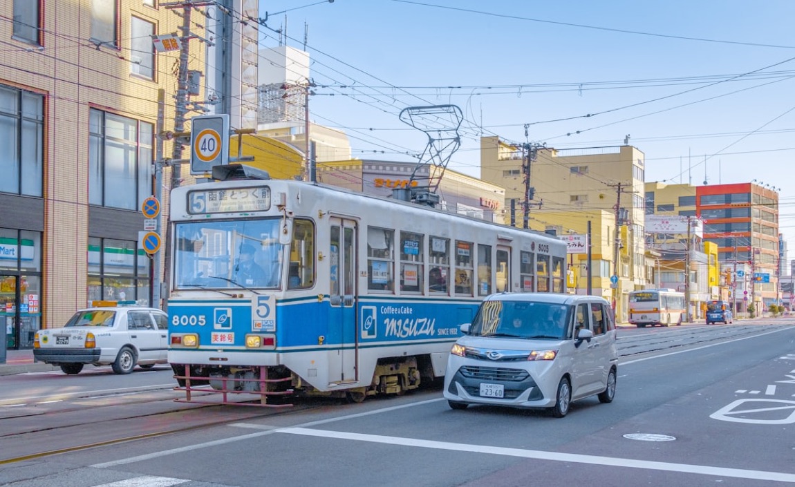 Un tram bianco e blu di Hakodate su una strada cittadina, con la destinazione in giapponese e i palazzi intorno