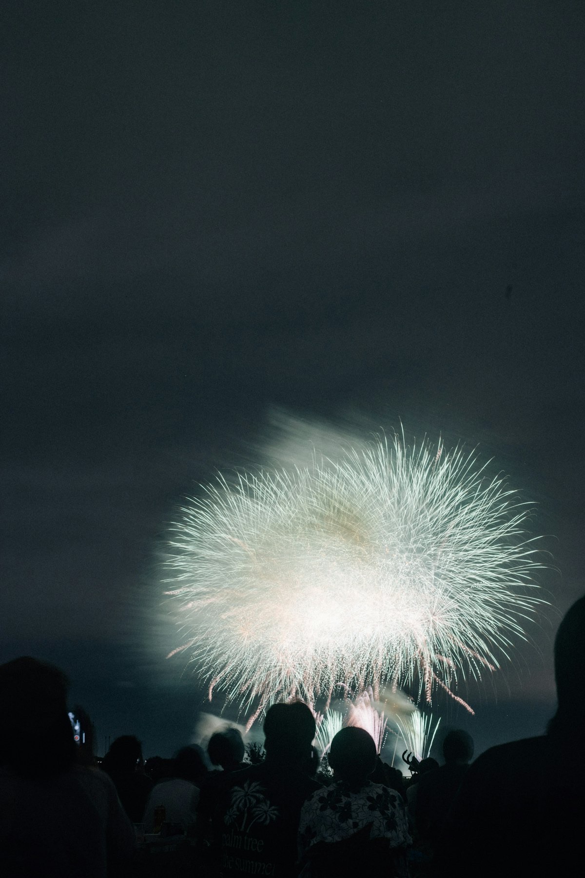 Un grande fiore di fuoco bianco esplode nel cielo notturno sopra la folla in silhouette a un festival estivo giapponese