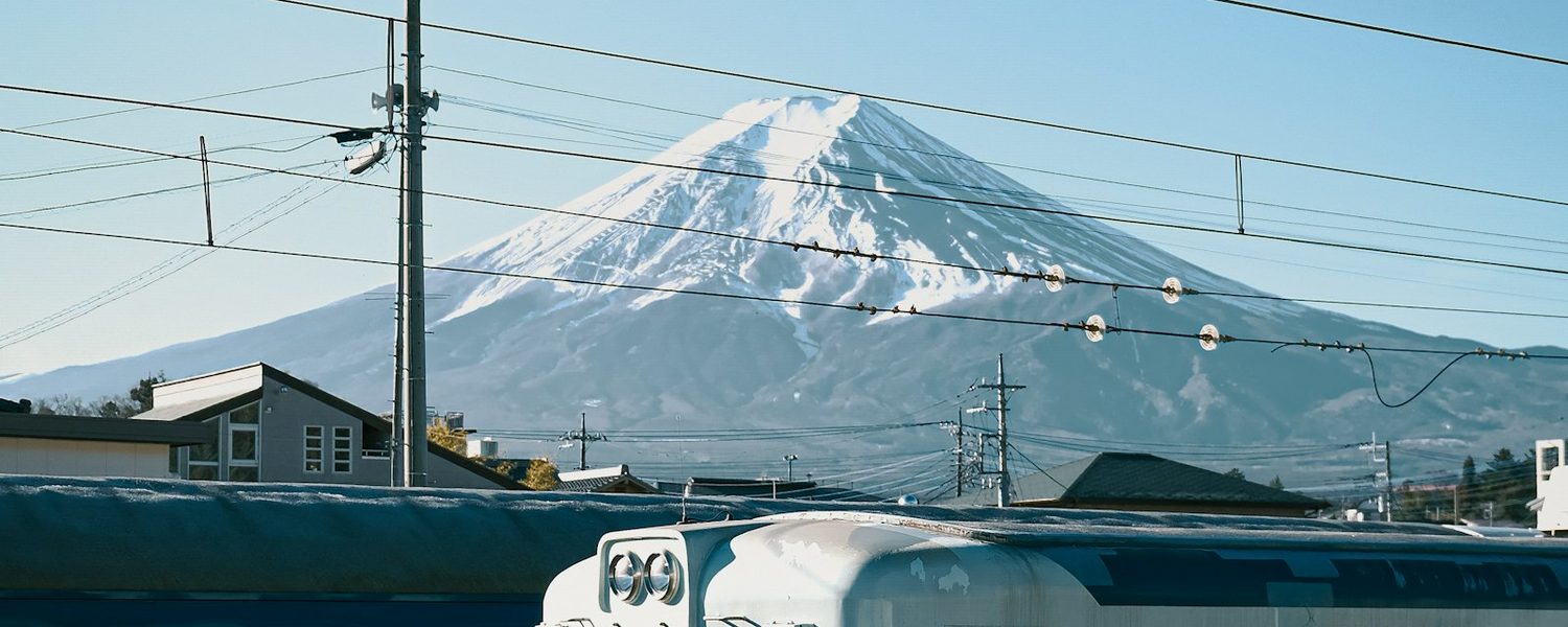 Il Monte Fuji innevato visto oltre i tetti, con un treno locale in primo piano