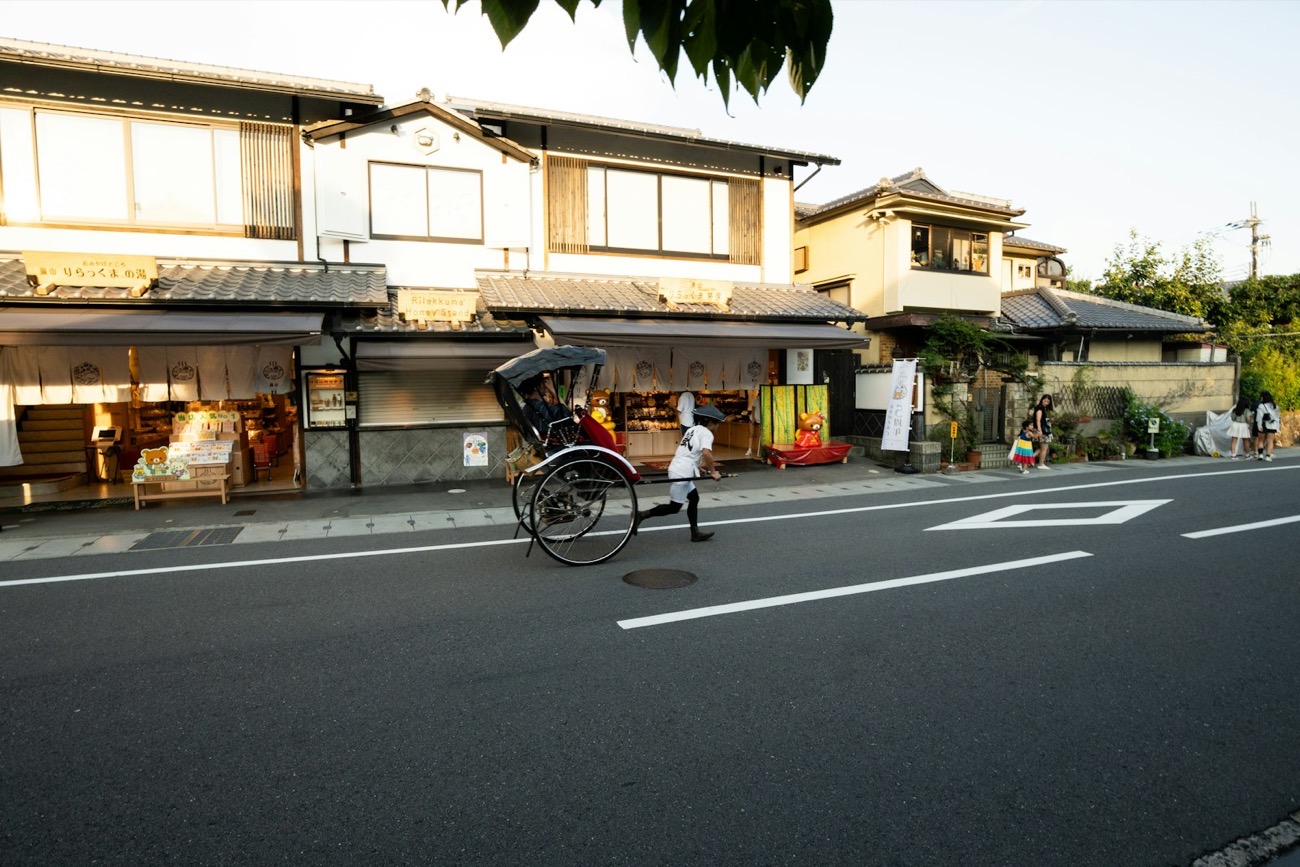 Un risciò tradizionale giapponese trainato da un conducente in divisa lungo una via di case e botteghe di legno a Kyoto, al tramonto
