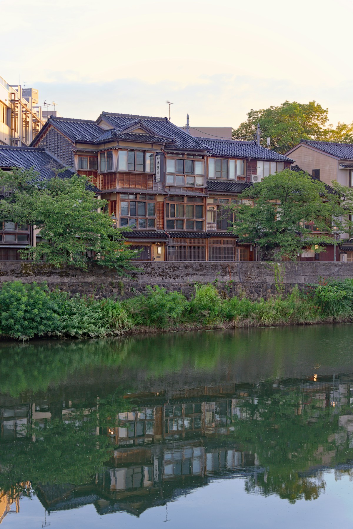 Un antico edificio tradizionale di Kanazawa riflesso nelle acque calme di un canale, circondato da alberi verdi al tramonto