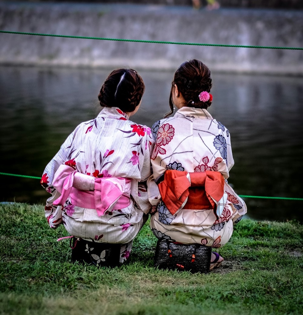 Due donne di spalle, in kimono a fiori con l'obi rosa e arancio annodato sulla schiena, sedute davanti all'acqua