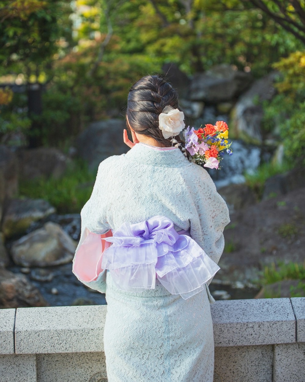 Una donna di spalle in kimono chiaro con un grande fiocco dell'obi lilla, davanti a un giardino con cascatella
