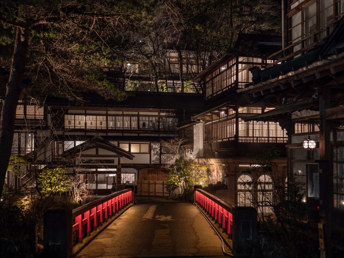Un antico ryokan di legno a più piani, illuminato di sera da lanterne calde, con un ponticello dalla ringhiera rossa