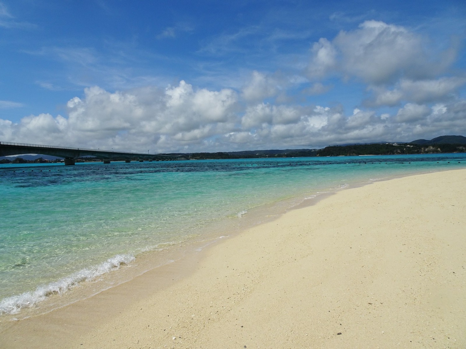 Il mare turchese di Okinawa, con la spiaggia di sabbia bianca e un ponte in lontananza sotto un cielo azzurro
