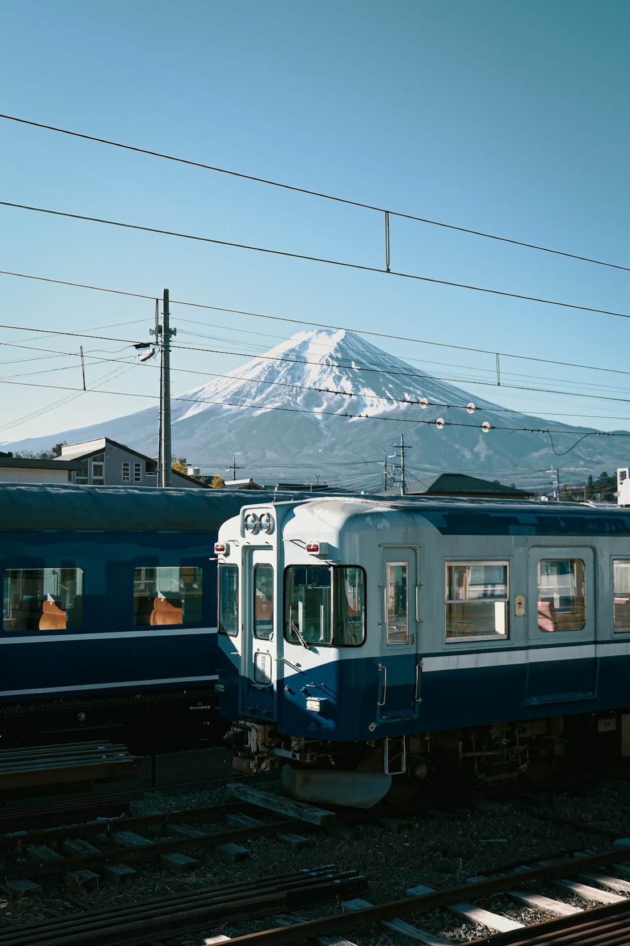 Un treno azzurro e crema fermo in stazione con il monte Fuji innevato sullo sfondo, sotto un cielo terso