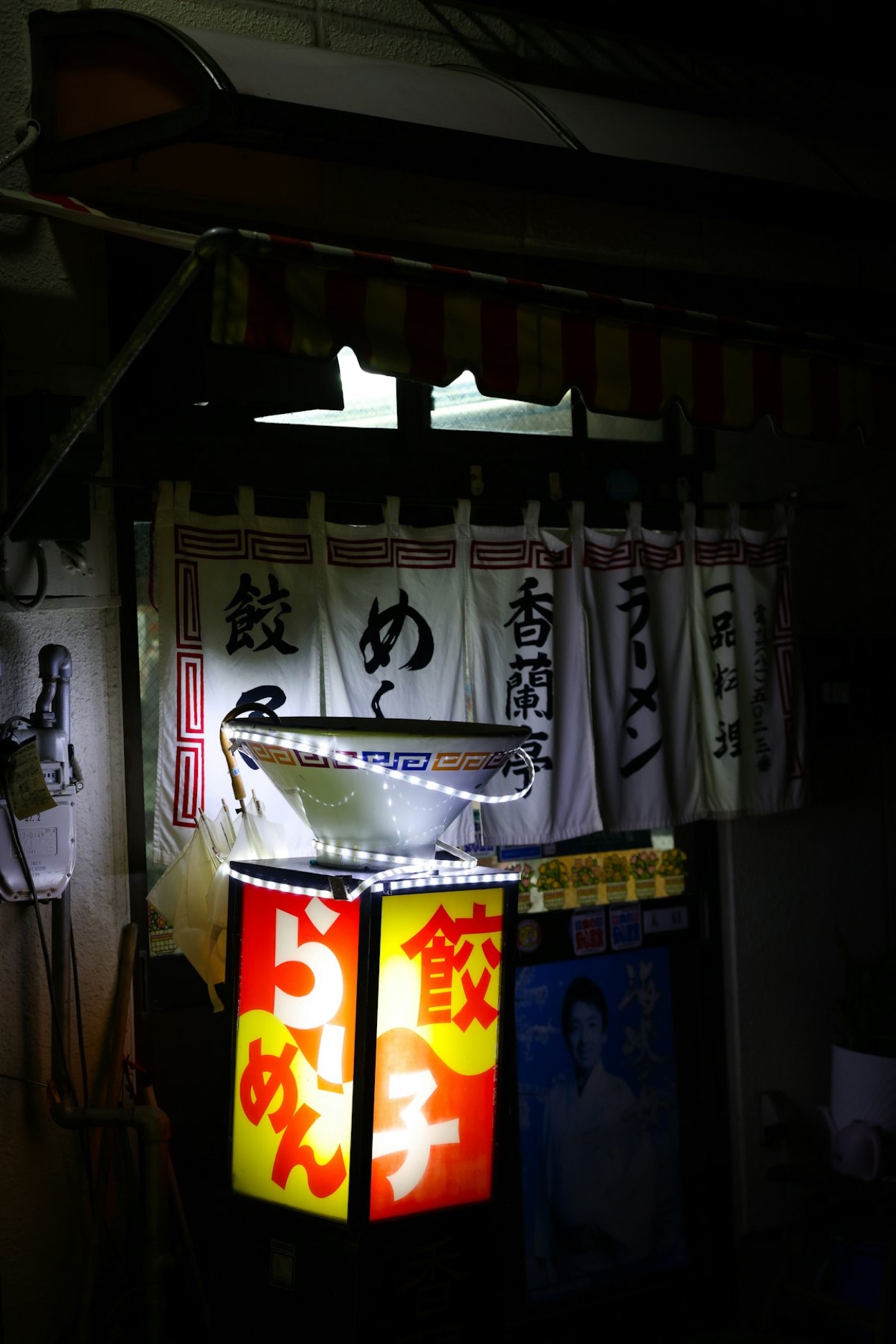 L'ingresso di un ramen-ya a Tokyo di sera, con il noren, l'insegna luminosa a forma di ciotola e le scritte "ramen" e "gyoza" in giapponese