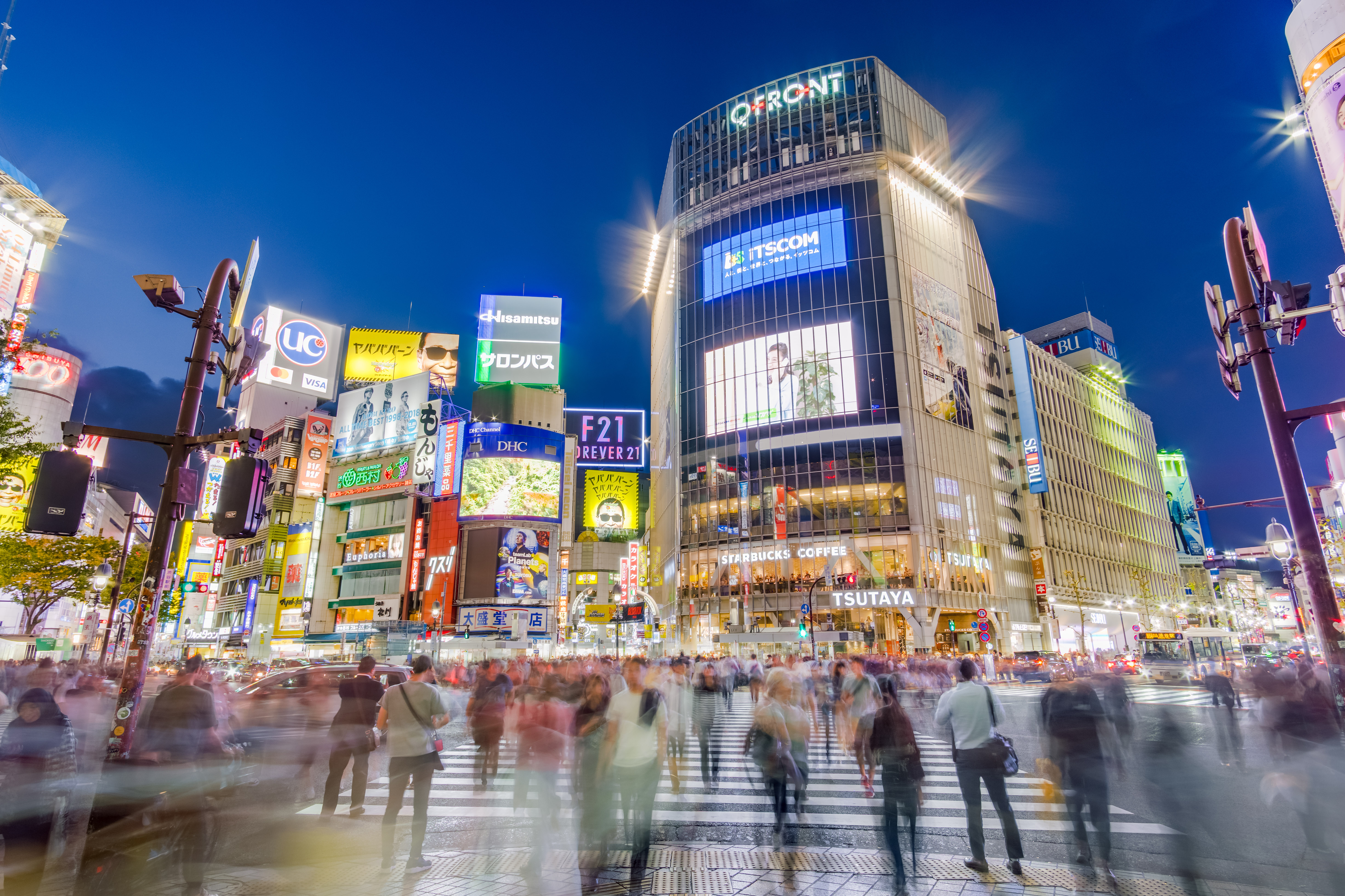 L'incrocio di Shibuya di notte, illuminato da insegne al neon e schermi giganti, con migliaia di persone che attraversano in tutte le direzioni