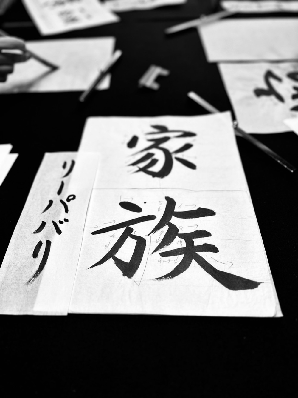 Fogli di calligrafia con i caratteri di "famiglia" (家族) in kanji e alcuni segni in katakana, su un tavolo scuro