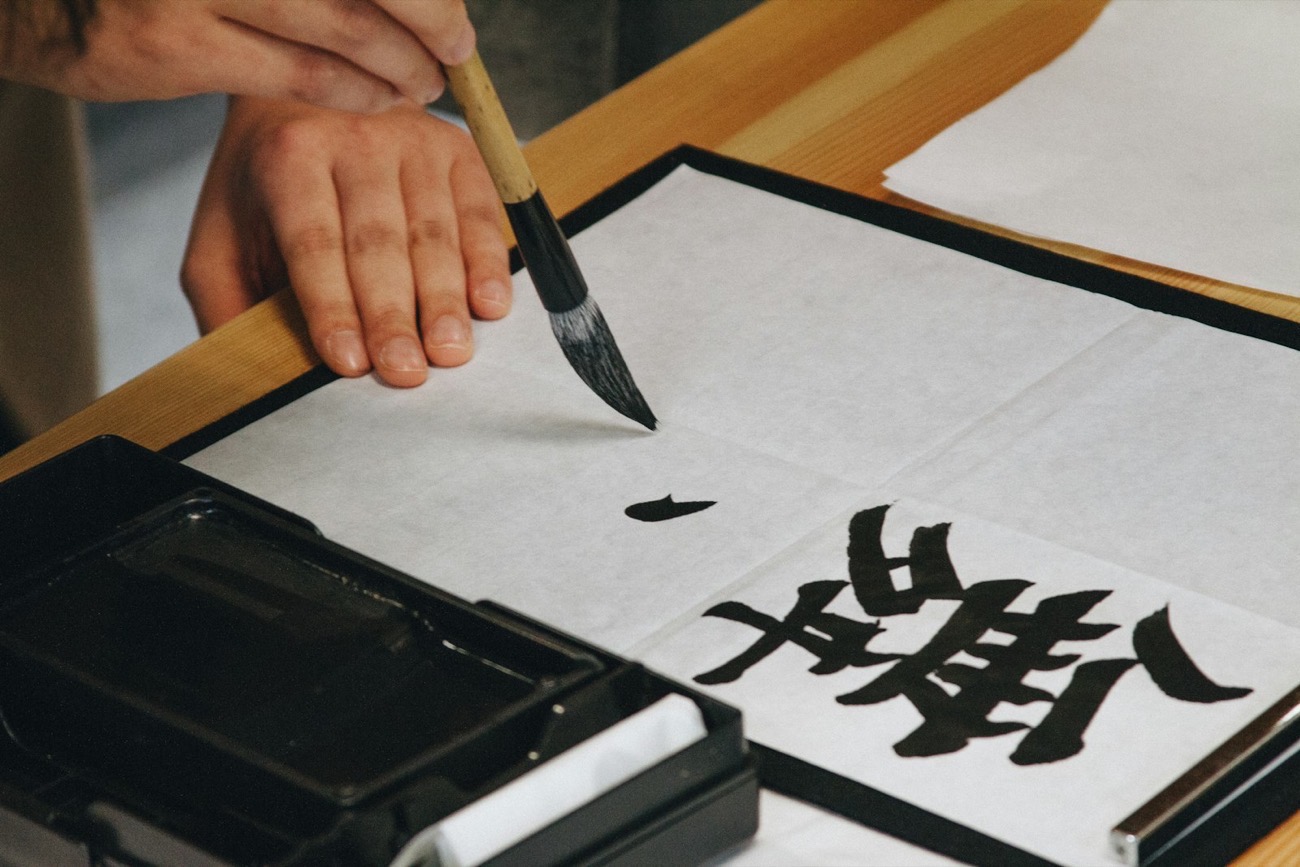 Una mano scrive un grande carattere kanji con un pennello nero su carta bianca, accanto a una vaschetta d'inchiostro nero