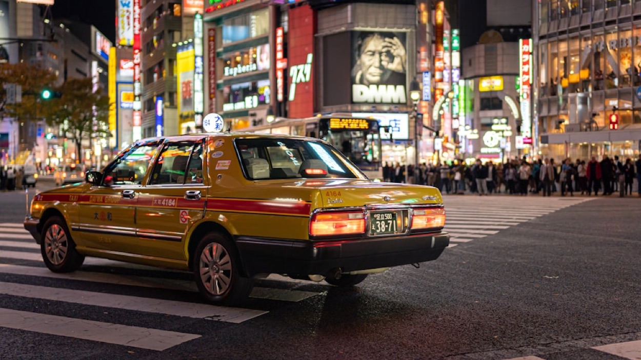 Un taxi giallo della compagnia Daiwa fermo a un incrocio di Shibuya di sera, con le insegne al neon dei palazzi sullo sfondo