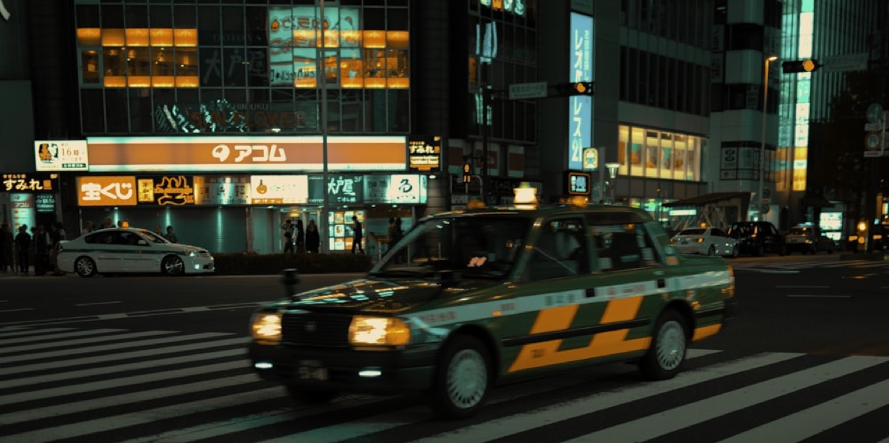 Un taxi giapponese verde e giallo attraversa un incrocio di notte, con le insegne luminose in giapponese sullo sfondo