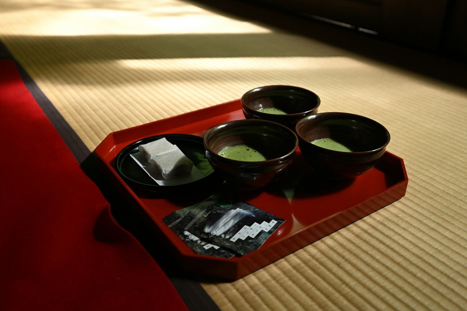 Un vassoio laccato rosso con tre ciotole di matcha appena montato e un dolcetto bianco, su un tatami attraversato dalla luce