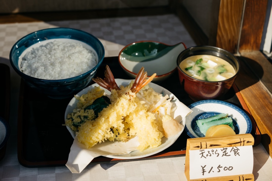 Un set di tempura giapponese: gamberi e verdure in pastella, riso, zuppa di miso e sottaceti su un vassoio, con un cartello in giapponese