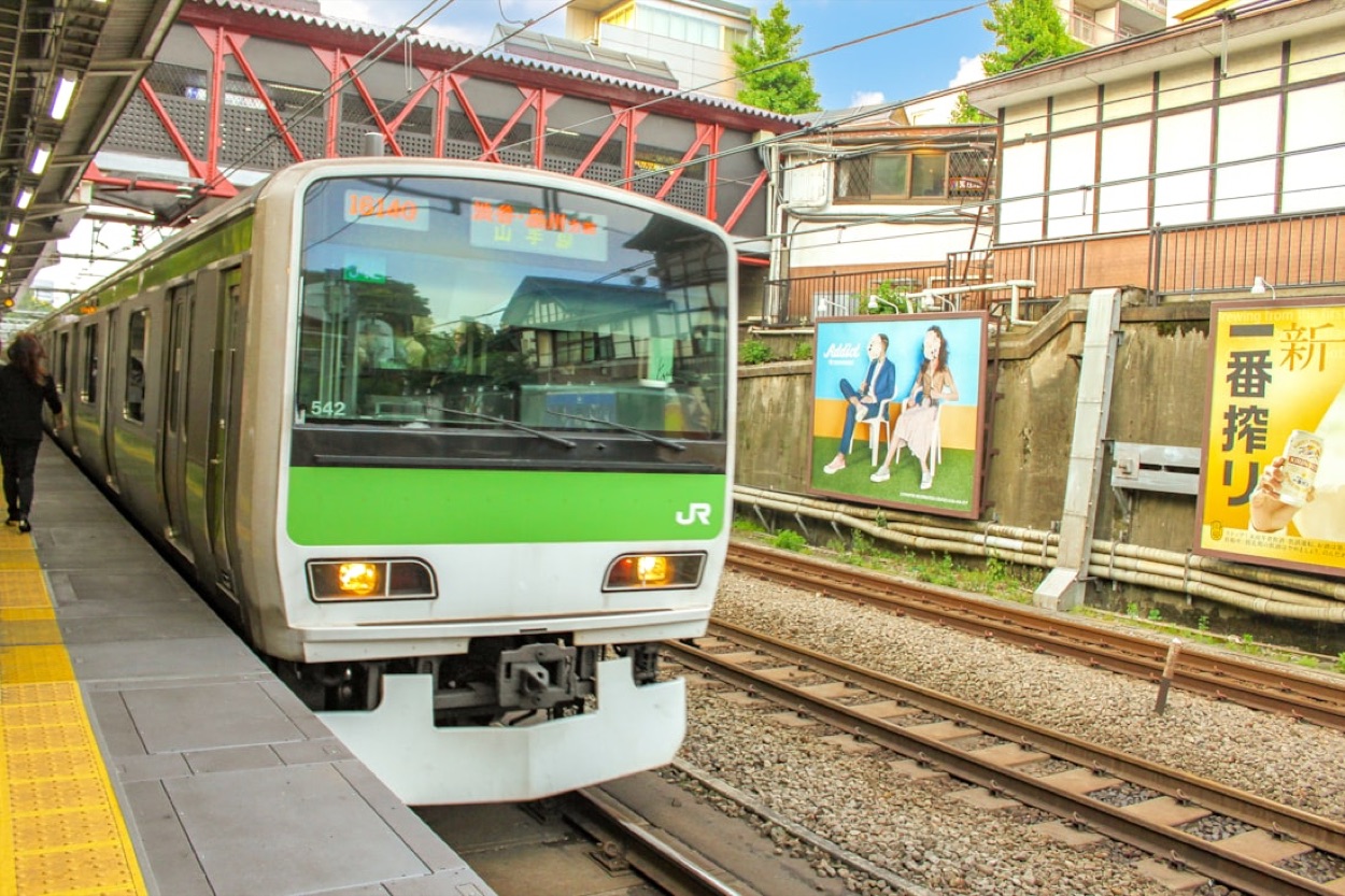 Un treno verde della linea Yamanote di Tokyo fermo al binario, con il logo JR e i cartelloni pubblicitari giapponesi