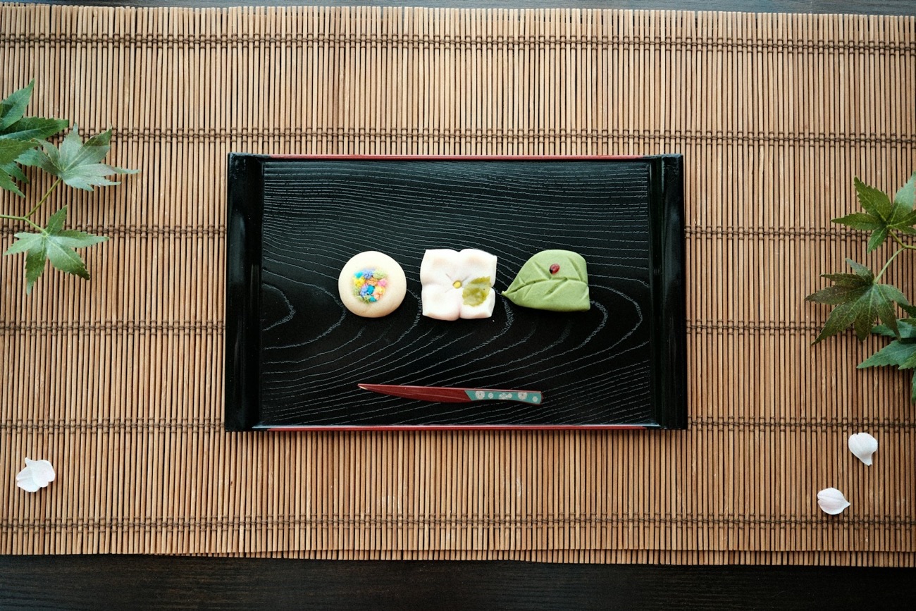 Tre wagashi di stagione — un fiore, una foglia d'acero e un dolcetto colorato — su un vassoio di lacca nera, con foglie e petali