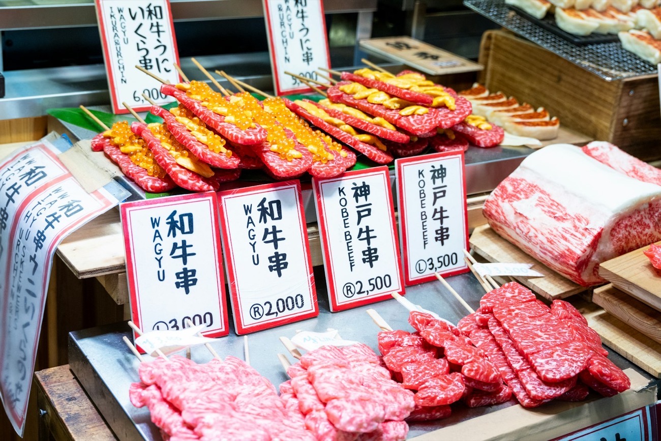 Un banco di mercato con spiedini di wagyu e manzo di Kobe dalla marezzatura intensa, con cartelli in giapponese e i prezzi in yen