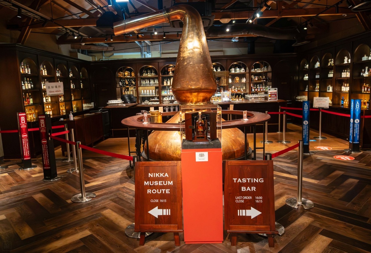Il museo della distilleria Nikka, con un grande alambicco di rame e gli scaffali pieni di bottiglie, e i cartelli "Nikka Museum Route" e "Tasting Bar"