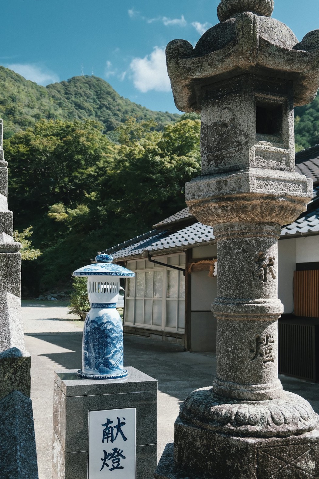 Una lanterna di porcellana bianca e blu di Arita accanto a un'antica lanterna di pietra, davanti a un santuario tra le montagne